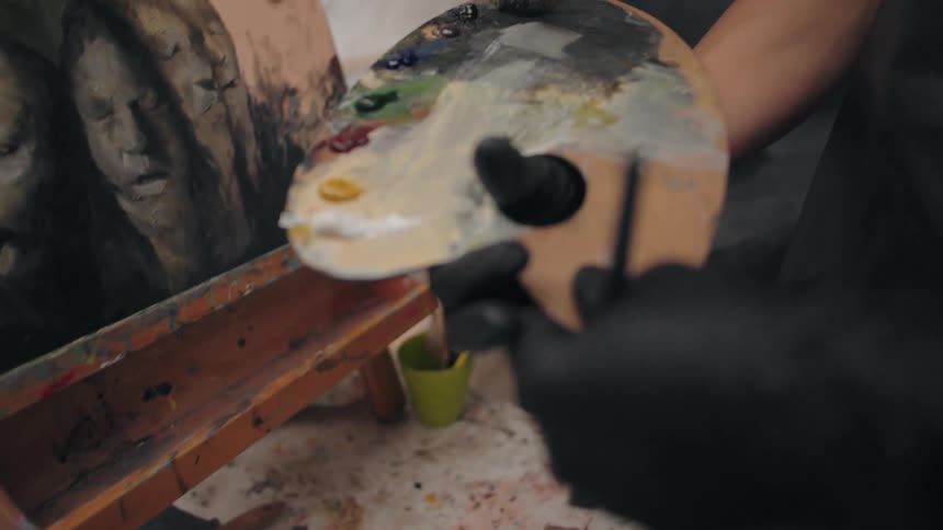 An artist in a black glove holding a wooden palette with various paint colors in their studio
