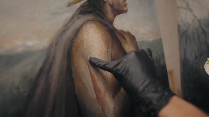 An artist wearing a black glove uses their thumb to gently blend and smudge paint on the arm of a figure in an oil portrait