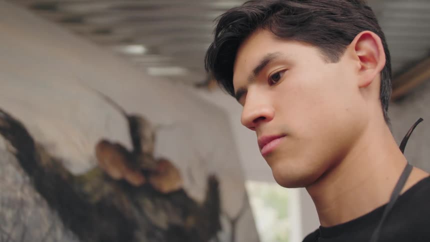 A closeup of a young mans face shows him concentrating and looking down at a large canvas that displays a blurred image while he stands in a studio with a metal roof and white walls