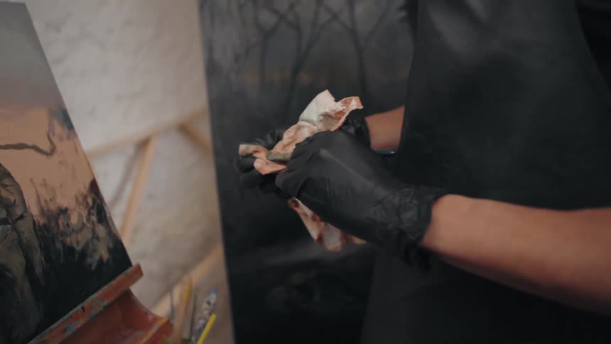 An artist in black gloves carefully wiping a paintbrush with a paint stained cloth in their studio