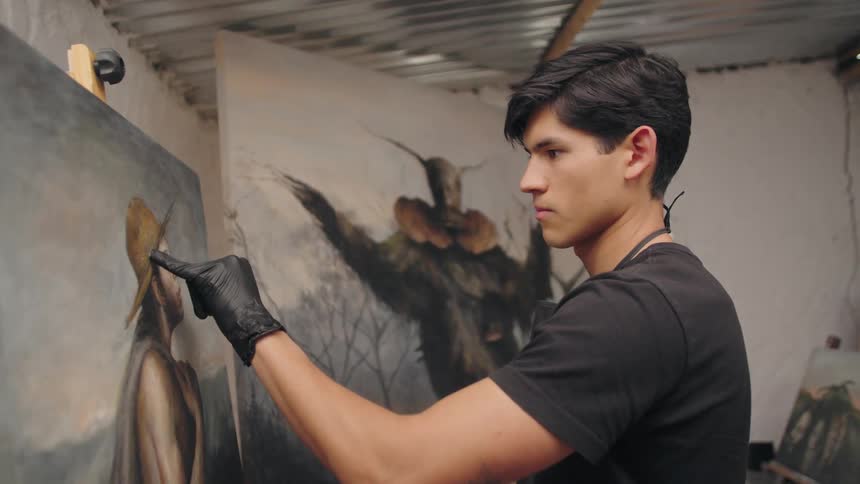 A focused young artist wearing a black glove carefully examines a painting on an easel in his studio