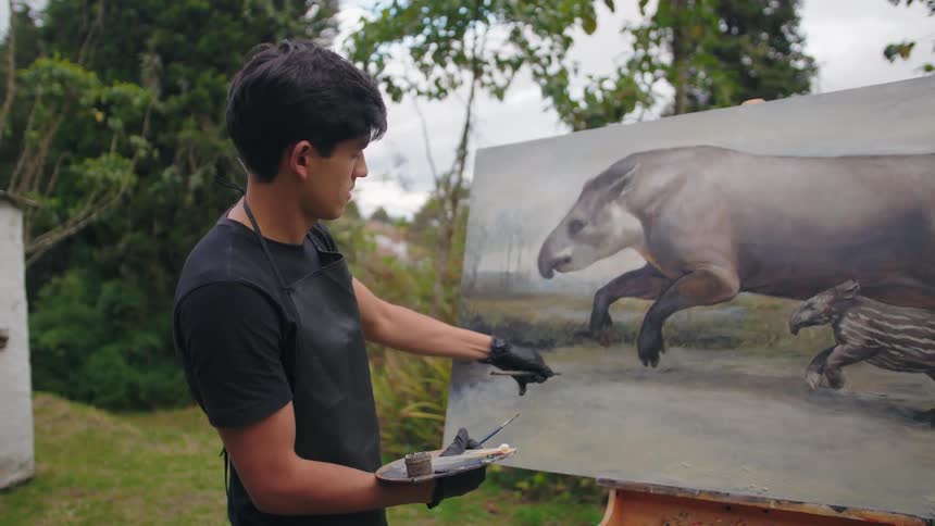 A focused young artist stands outdoors in a garden painting a large realistic depiction of a tapir and its calf on an easel