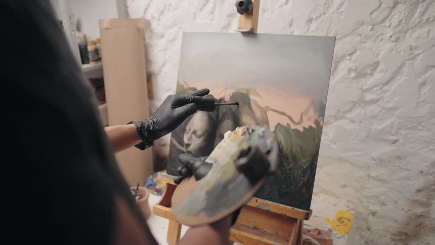 A closeup of an artist painting on a canvas with a palette in hand