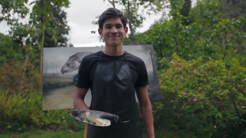 Young artist smiles holding a paint palette in front of his outdoor painting