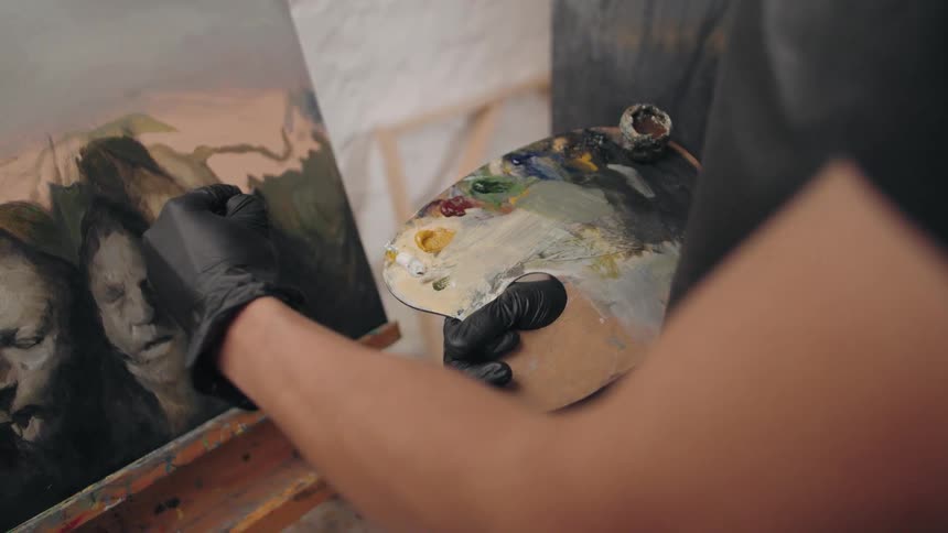 A person wearing black gloves holds a wooden palette in the right hand and uses a paintbrush in the left hand to apply paint to a canvas showing dark stylized figures