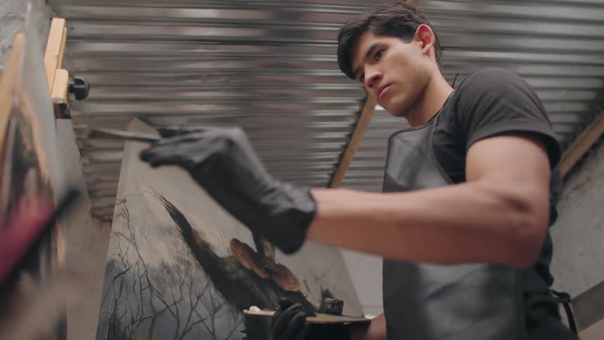 A focused young male artist in a black apron and gloves works intently on a large moody painting in his studio under a corrugated metal roof