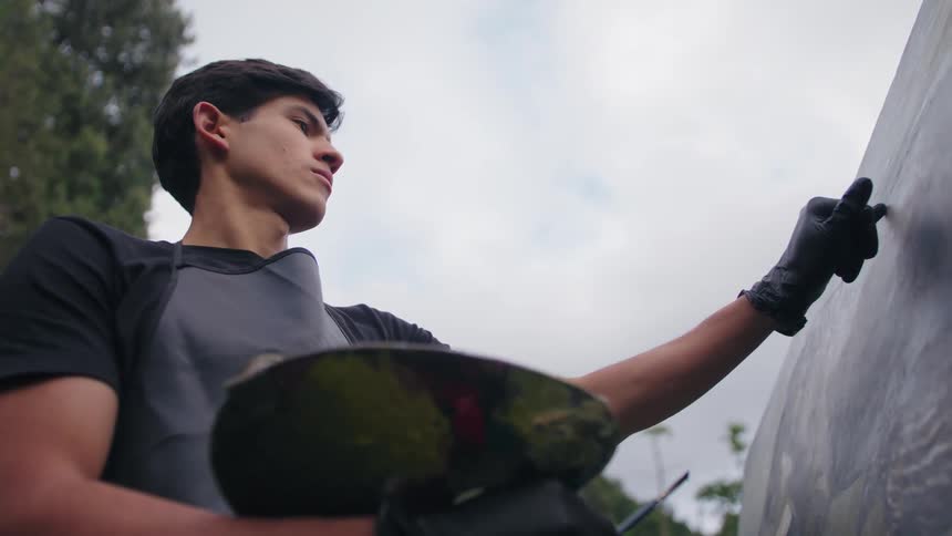 A man wearing black gloves and an apron holds a palette while touching a large canvas with his hand looking intently at the artwork outdoors beneath an overcast sky and green trees