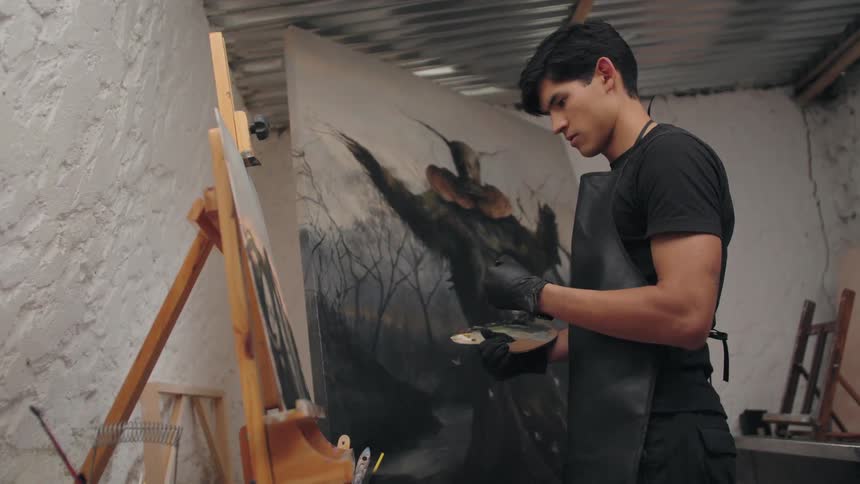 A man wearing black gloves and an apron stands in an art studio with a white wall and metal roof looking intently at a large canvas on an easel and applying paint with a brush