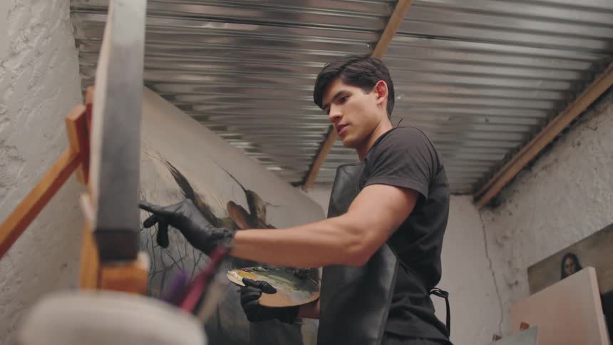 The video shows a focused male artist wearing a black shirt a black apron and black gloves intently working on a large canvas secured to an easel he is holding a palette and using a brush to apply paint to a dark textured image inside what appears to