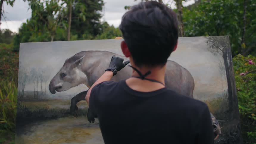 A man wearing black gloves and a black tshirt stands with his back to the camera outdoors among green trees painting a large canvas
