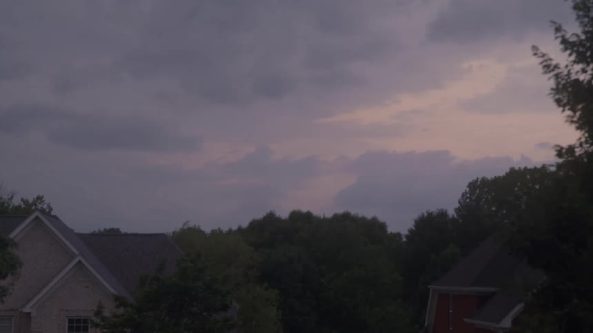 Dark heavy clouds move slowly across the sky over a dense line of treetop foliage with the rooftops of suburban houses visible below