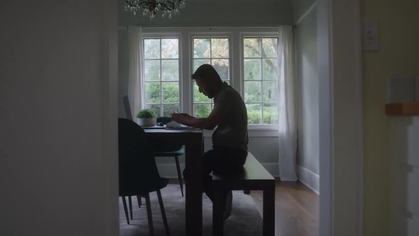 A shot looks down a doorway into a brightly lit kitchen where a person is sitting at a dining table working or reading the scene is welllit by natural light coming through the windows at the far end of the room