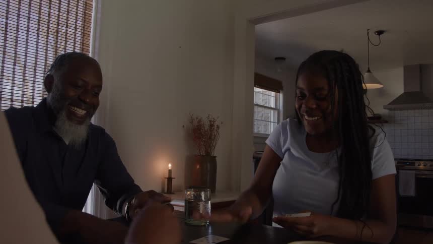 A man a young black woman and another person sit together at a dining table at midday smiling and enjoying a lively card game