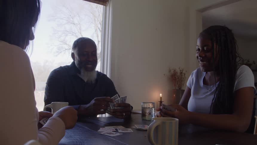 Three people sit around a table and play a card game handling cards and smiling at each other in a room with a large sunlit window