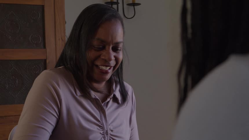 A middleaged woman smiles and laughs during a conversation with an unseen person in a welllit indoor setting she is wearing a light purple top and the background features a wooden cabinet and an ornamental wall sconce