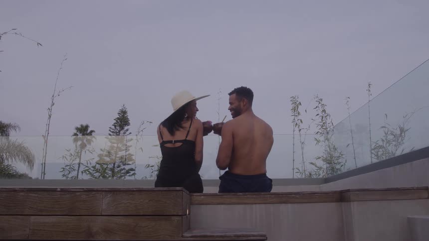 A smiling couple shares coconut drinks by a pool under a twilight sky