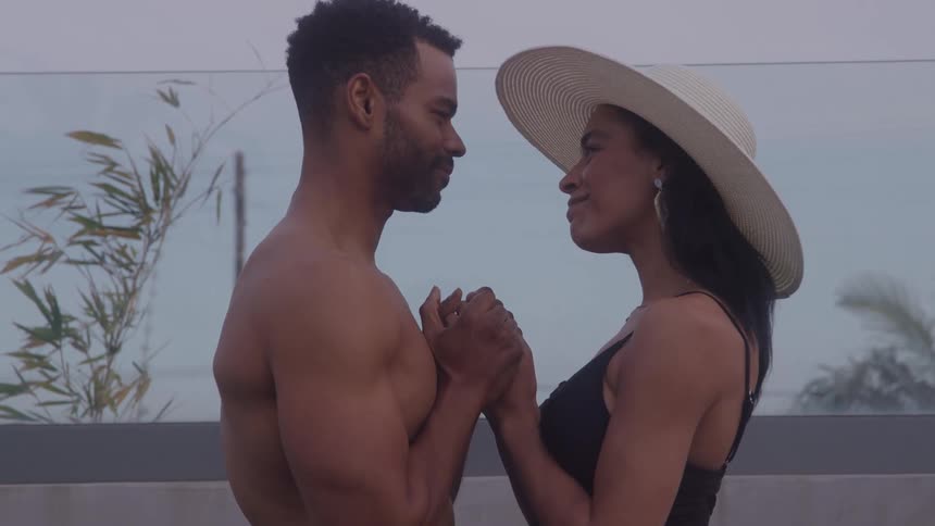 A side profile of an attractive couple holding hands and looking into each others eyes in an outdoor setting the woman is wearing a dark swimsuit and a widebrimmed white sun hat while the shirtless man smiles at her