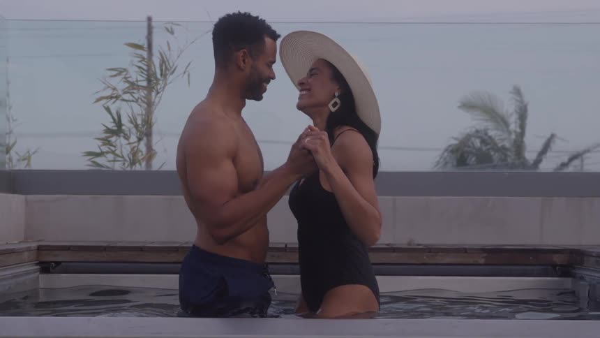 A man and a woman are standing facetoface in a hot tub both smiling and holding hands the woman is wearing a black swimsuit and a widebrimmed sun hat while the man is shirtless