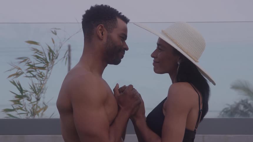 A couple stands facing each other at a public pool area under the midday sun holding hands the man is shirtless and the woman wears a swimsuit and a sun hat both enjoying a lighthearted moment together