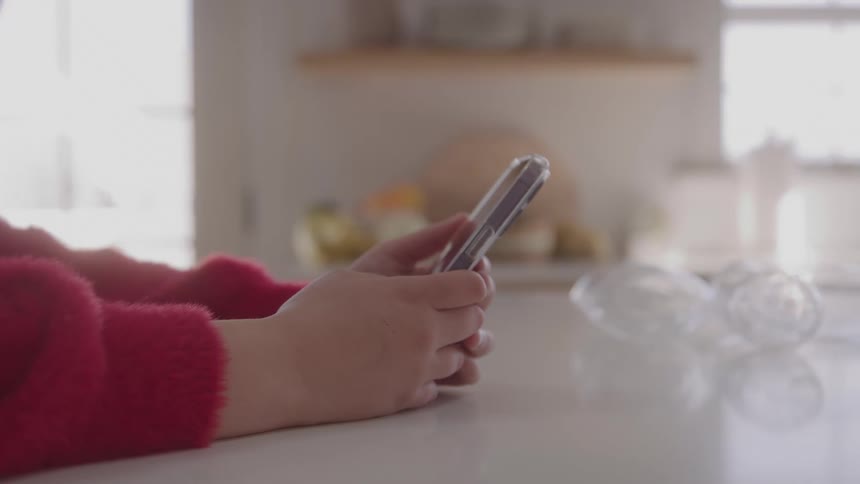 Hands holding a smartphone rest on a white countertop the person wears a red sweater and a kitchen counter and cabinets are blurred in the background