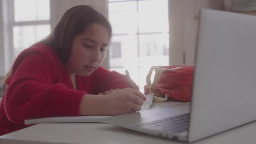 A young girl writes in a notebook beside her laptop at a kitchen counter