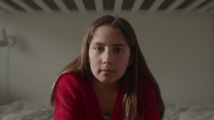 A young girl looks into the camera while sitting in a softly lit bedroom