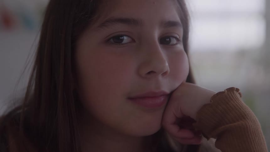 A young girl smiles softly into the camera resting on her hand indoors