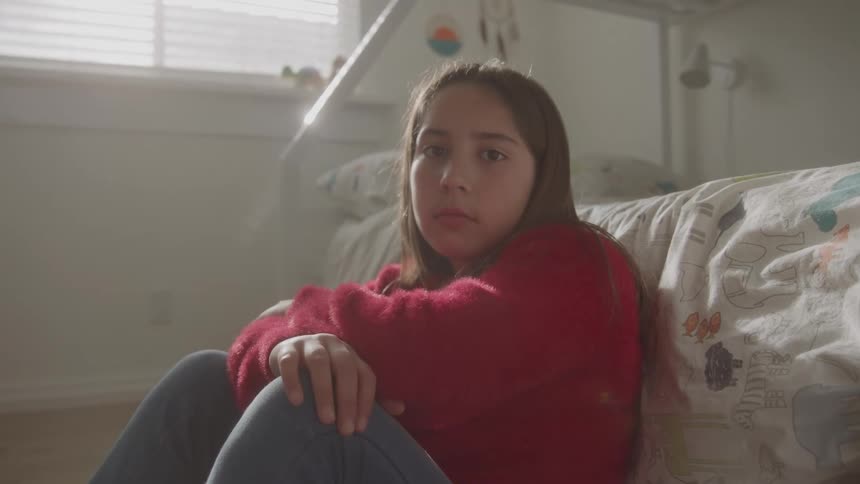 Girl sitting on the floor leaning against the bed in a naturally lit bedroom