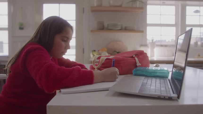 A young girl writes in a notebook beside her laptop at a kitchen counter