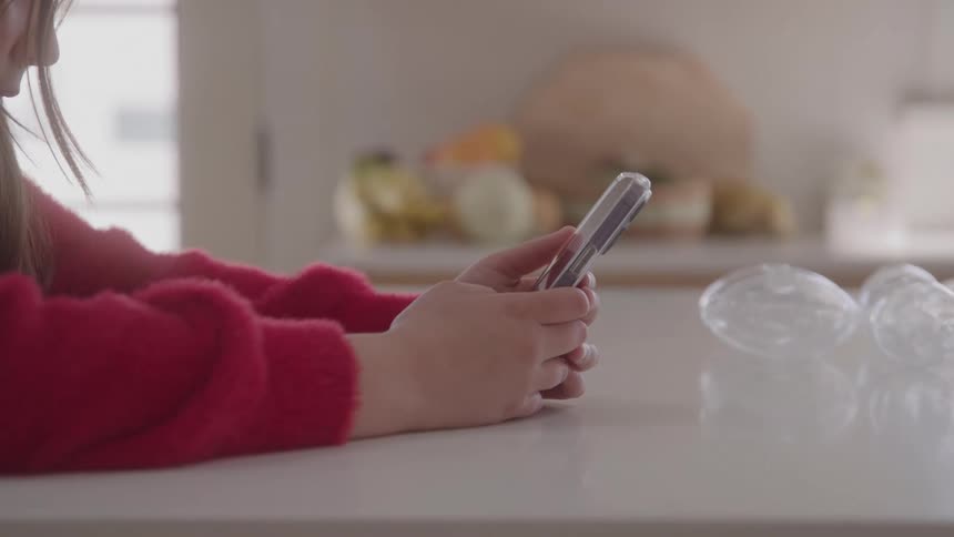 A young person in a red sweater is sitting at a white kitchen counter looking down at a smartphone they hold in their hands in the background there are various outoffocus kitchen items including a bowl of fruit