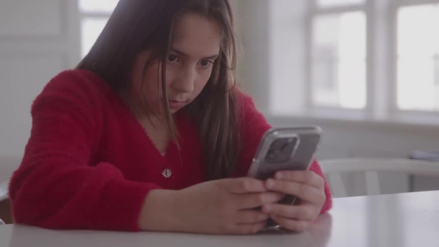 A young person in a red sweater is sitting indoors focusing intently on their smartphone they have a serious or concerned expression as they look down at the screen