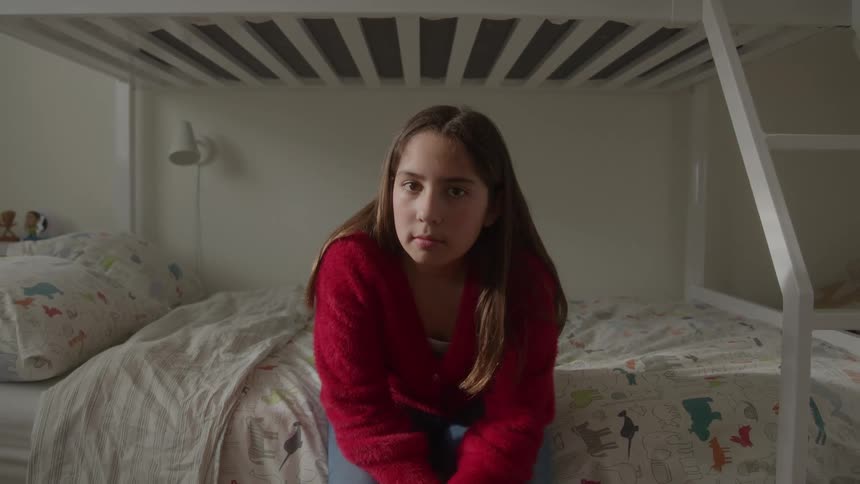 A young girl wearing a red sweater sits centered on the lower bunk bed in a brightly lit bedroom turning her head from the side to look directly into the camera