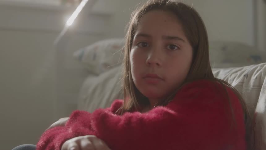 A teenage girl sitting leaning against a bed with a serious and melancholic expression