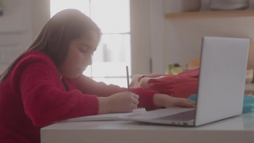 A young girl writes in a notebook at her desk with a laptop