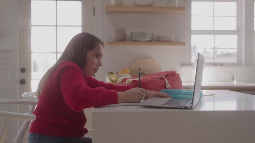 A young girl studies and writes beside her laptop at the kitchen counter