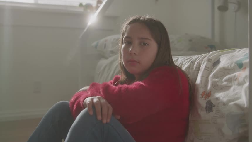 The girl is sitting on the floor by her bed