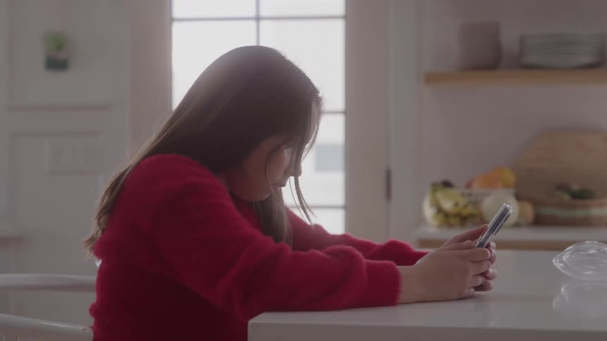 A young girl looks down at her phone while sitting at a kitchen counter