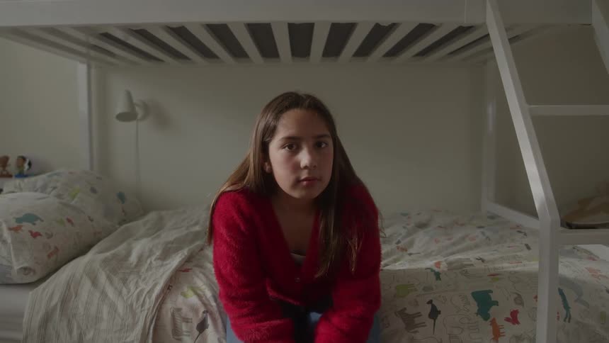 A young girl in a red sweater sits on a bunk bed gazing directly at the camera