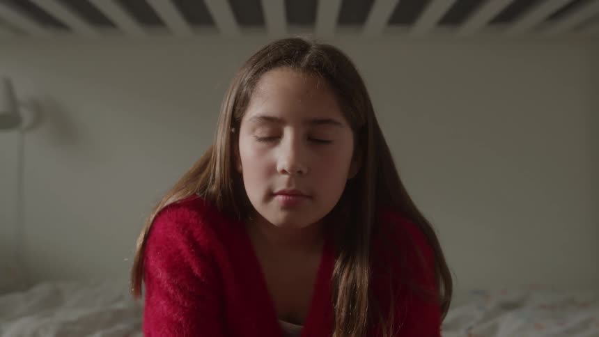 A young girl wearing a red cardigan sits on a bed looking directly at the camera with a serious contemplative expression the background is a dimly lit bedroom with the slats of a bunk bed visible above her head