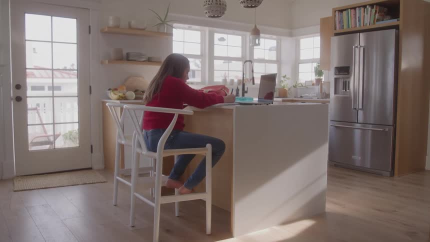 A wide shot shows a bright modern kitchen with a girl in a red sweater sitting at an island counter she is facing a laptop placed on the counter in front of her