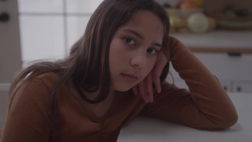 A young girl sits at a table resting on her hand in a softly lit kitchen
