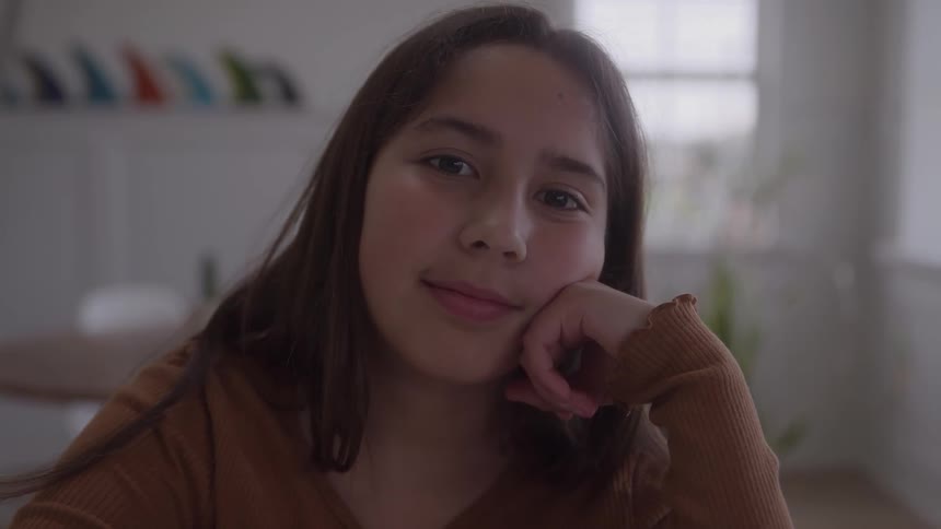 A medium closeup shows a young woman with long brown hair resting her chin on her hand looking directly at the camera she maintains a soft slightly contemplative expression in a welllit blurred indoor environment