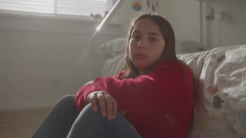 A young girl sits on the floor with her arms crossed and her legs bent turns her head slowly and looks directly forward with a somber expression in a brightly lit bedroom