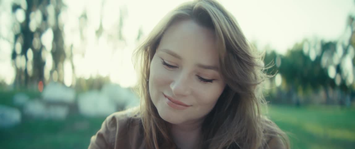 Woman with light brown hair sits in grassy area with trees in the background under soft light