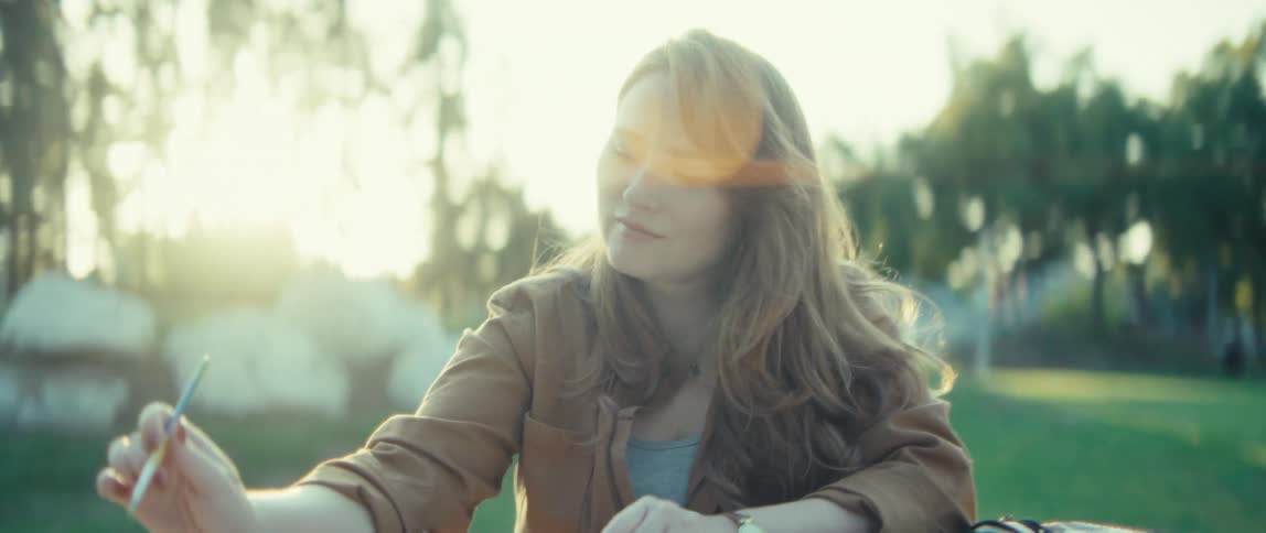A young woman sits outdoors at a picnic table actively drawing or painting in a sketchbook her long wavy hair is loose and the background consists of blurred sunlit greenery