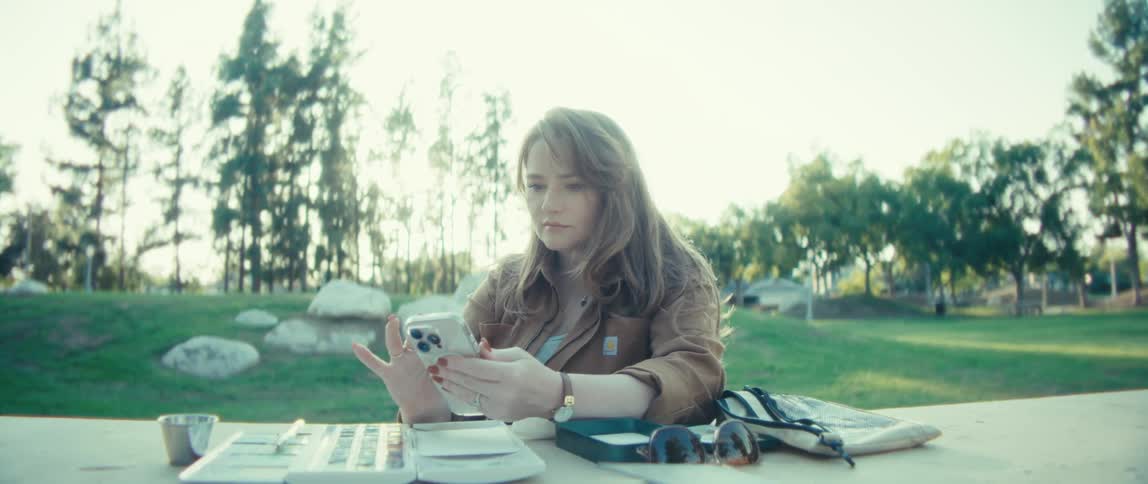 A woman sits outdoors at a picnic table on a sunny day looking down and interacting with her smartphone there are art supplies including a small palette and box scattered on the table beside her