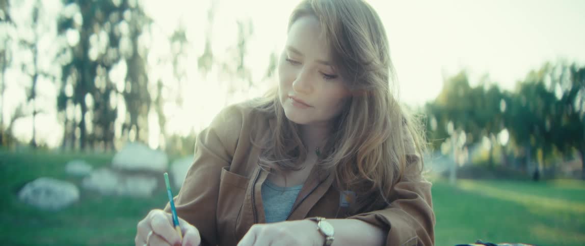 A young woman is sitting outside at a table focused on writing or sketching with a pencil she wears a brown jacket and is outdoors on a sunny day with trees in the background