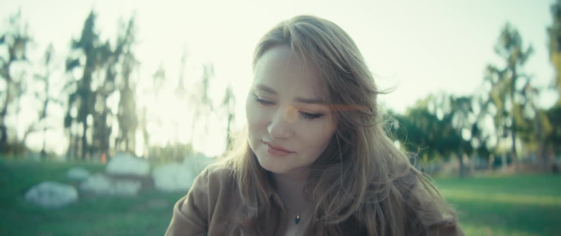 A closeup shot focuses on a young woman outdoors in a park with sunlight illuminating her long wavy hair she looks up with a serene expression then gently lowers her gaze