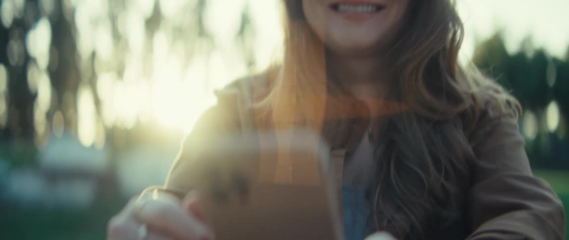 A young woman sits outside in natural light with her long wavy hair framing her face she looks down and then slightly up with a gentle soft smile