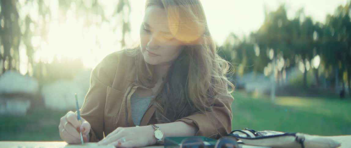 A young female artist sits on a bench in a public park at midday painting thoughtfully on a piece of paper as sunlight filters through the trees